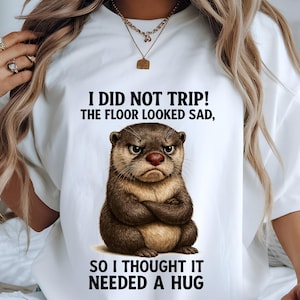 May include: White t-shirt with a cartoon otter illustration. The otter has a stern expression and crossed arms. The text on the shirt says, "I DID NOT TRIP! THE FLOOR LOOKED SAD, SO I THOUGHT IT NEEDED A HUG."