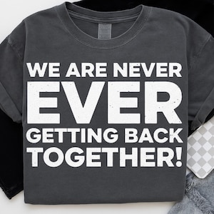 May include: Dark gray t-shirt with the white text "WE ARE NEVER EVER GETTING BACK TOGETHER!". The shirt has a crew neck and short sleeves. The text is in a bold, sans-serif font.
