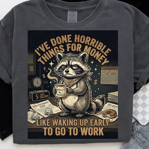 Puede incluir: Camiseta gris oscuro con un mapache sosteniendo una taza de café. El texto en la camiseta dice "He hecho cosas horribles por dinero" y "Como despertarse temprano para ir a trabajar." La taza dice "World's No. 1 Worker."