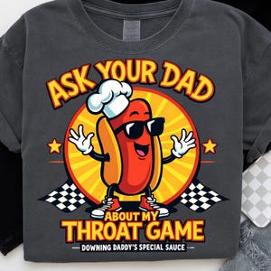 May include: Dark gray t-shirt featuring a cartoon hot dog wearing sunglasses and a chef's hat. The text reads "ASK YOUR DAD ABOUT MY THROAT GAME - DOWNING DADDY'S SPECIAL SAUCE" in yellow and red.