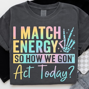 May include: Dark gray t-shirt with the text "I MATCH ENERGY SO HOW WE GON' Act Today?" in a rainbow gradient. A skeleton hand makes a peace sign. The shirt is a casual wear item.