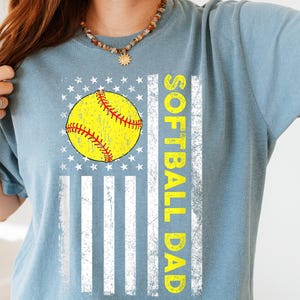 May include: A light blue t-shirt featuring a softball design. The design includes a yellow softball with red stitching, set against a white and blue flag graphic. The words "SOFTBALL DAD" are printed vertically in yellow.