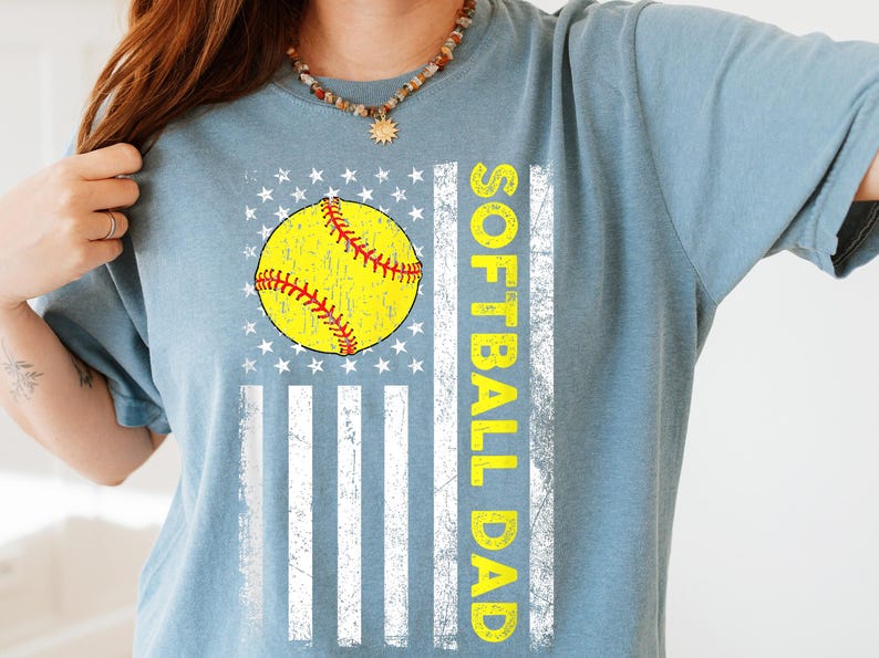 May include: A light blue t-shirt featuring a softball design. The design includes a yellow softball with red stitching, set against a white and blue flag graphic. The words "SOFTBALL DAD" are printed vertically in yellow.