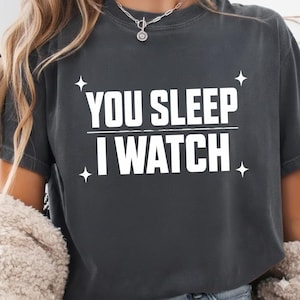 May include: Dark gray t-shirt with the white text "YOU SLEEP I WATCH" and four white starburst graphics. The shirt has a crew neck and short sleeves.