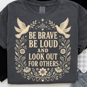 May include: A charcoal gray t-shirt featuring a cream-colored graphic print of two doves, floral vines, and the text: BE BRAVE BE LOUD AND LOOK OUT FOR OTHERS. The design is centered on the chest, framed by symmetrical leaves and flowers.
