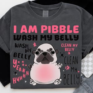 Pode incluir: Camiseta cinza escuro com o texto "I AM PIBBLE WASH MY BELLY" em rosa e preto. Um desenho de pug com uma barriga rosa está centralizado abaixo do texto. O texto adicional inclui "WASH MY BELLY" e "CLEAN MY BELLY".