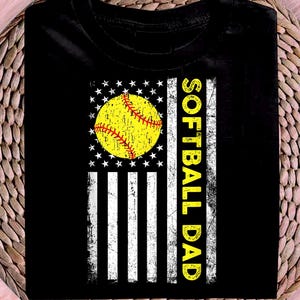 May include: Black t-shirt featuring a softball design. The design incorporates a yellow softball with red stitching, set against a white star-studded circle and white stripes, resembling a flag. The words "SOFTBALL DAD" are printed vertically in yellow.