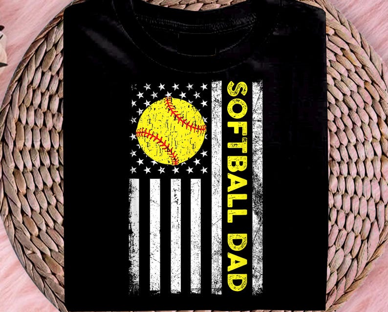 May include: Black t-shirt featuring a softball design. The design incorporates a yellow softball with red stitching, set against a white star-studded circle and white stripes, resembling a flag. The words "SOFTBALL DAD" are printed vertically in yellow.
