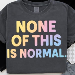 May include: Dark grey crew neck sweatshirt featuring the phrase "NONE OF THIS IS NORMAL" in pastel rainbow lettering. The sweatshirt is a solid colour and has a classic, casual style.