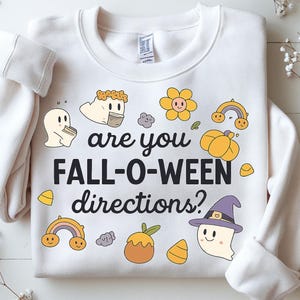 Könnte beinhalten: Weißes Sweatshirt mit dem schwarzen Schriftzug "are you FALL-O-WEEN directions?". Das Design umfasst Cartoon-Geister, Kürbisse, Regenbogen und andere herbstliche Grafiken in Gelb, Orange und Lila.
