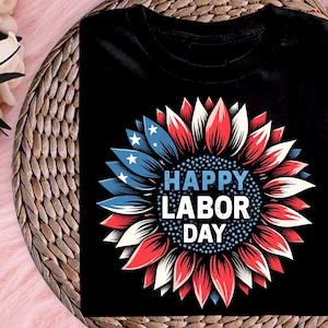 Könnte beinhalten: Schwarzes T-Shirt mit einem Sonnenblumen-Design in Rot, Weiß und Blau und dem Text "HAPPY LABOR DAY". Die Blütenblätter sind kreisförmig angeordnet, mit blauen Sternen. Das T-Shirt ist aus weichem Material.