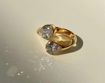 Könnte beinhalten: Ein goldener Ring mit zwei tropfenförmigen, klaren Steinen. Der Ring ist offen und kann an verschiedene Fingergrößen angepasst werden.