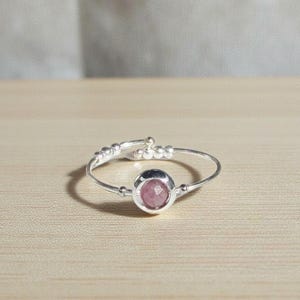 May include: A silver ring with a round, pink gemstone at the center. The band has a delicate design with small decorative elements. The ring is set against a light wooden surface.