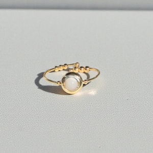 Puede incluir: Un delicado anillo dorado con una pequeña piedra preciosa blanca y redonda en el centro. La banda del anillo es fina y presenta pequeñas cuentas decorativas. El anillo se encuentra sobre un fondo blanco liso, resaltando su simple elegancia.