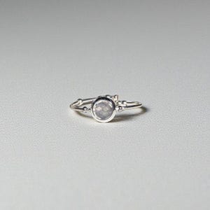 May include: A delicate silver ring with a round, light-colored gemstone at the center. The ring has a simple band and small decorative elements on either side of the gemstone. The ring is set against a plain, light-colored background.