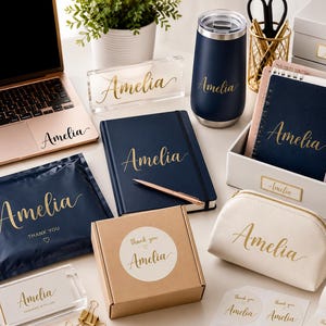 May include: A collection of personalized items featuring the name Amelia in gold script, including a navy notebook, a navy tumbler, a white pouch, a laptop decal, a brown cardboard box, a clear acrylic block, and various stickers and labels.