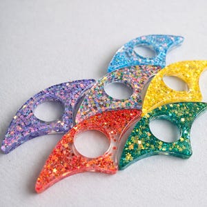 May include: A collection of colorful, glitter-filled thumb page holders. The holders are in various colors, including purple, blue, red, yellow, and green. Each holder has a circular opening for the thumb and is shaped to hold a book open. The glitter is star-shaped.