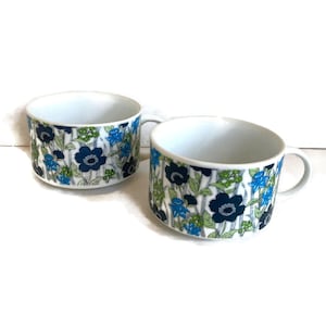 May include: Two white ceramic mugs with a floral pattern in shades of blue, green, and black. The mugs have a wide, rounded shape and a handle on the right side.