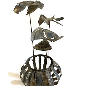 May include: A metal sculpture of a flower arrangement in a vase. The flowers are made of recycled metal and have a rustic, weathered look. The vase is also made of metal and has a geometric design.