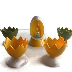 May include: A collection of egg-shaped kitchen items. Includes two yellow egg cups, two green egg cups, and a yellow and white egg timer. The egg cups have a jagged edge design and sit on white bases.