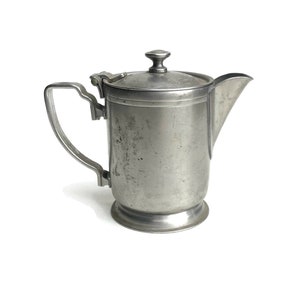 May include: A silver metal creamer or pitcher with a lid and a handle. The creamer has a slightly worn, aged look.