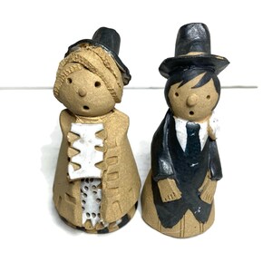 May include: Two ceramic figurines of a bride and groom. The bride wears a tan dress and black hat. The groom is in a black suit and top hat. Both have simple facial features and are on a white background.