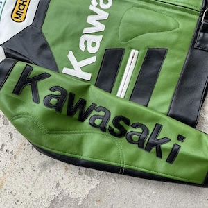 K&ouml;nnte beinhalten: Gr&uuml;ne und schwarze Motorradjacke mit dem Wort "Kawasaki" in wei&szlig;en und schwarzen Buchstaben. Die Jacke hat einen wei&szlig;en und schwarzen Aufn&auml;her mit dem Wort "Michelin". Die Jacke ist aus Leder.
