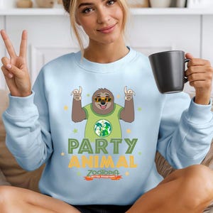 A woman wearing a light blue sweatshirt with a cartoon sloth graphic and the text "PARTY ANIMAL" is sitting on a couch and holding a black mug.
