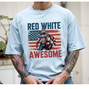 May include: A light blue t-shirt featuring a graphic of Captain America standing in front of a distressed American flag. The text "RED WHITE" appears in dark blue above the character, and the word "AWESOME" is printed in red below the graphic.
