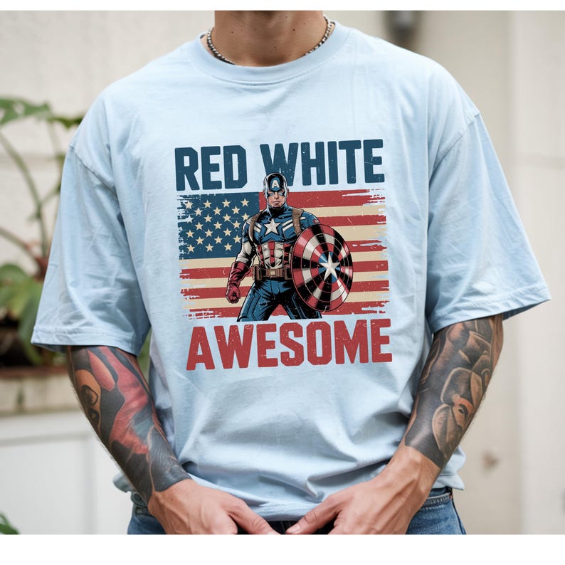 May include: A light blue t-shirt featuring a graphic of Captain America standing in front of a distressed American flag. The text "RED WHITE" appears in dark blue above the character, and the word "AWESOME" is printed in red below the graphic.