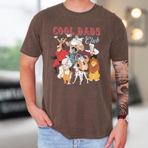 A man wearing a brown t-shirt with a graphic design of cartoon characters from the animated film "Cool Dads Club".