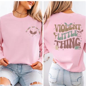 May include: A light pink sweatshirt with the text "VIOLENT LITTLE THING" in a retro font. The front of the sweatshirt has a dragon graphic and text. The back of the sweatshirt has a larger graphic with dragons and stars.