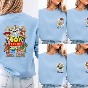 May include: Light blue sweatshirt featuring Toy Story characters. The back of the sweatshirt has a large graphic with Woody, Buzz Lightyear, and other characters, with "Toy Story" and "Est. 1995" text. Smaller character graphics are on the front.