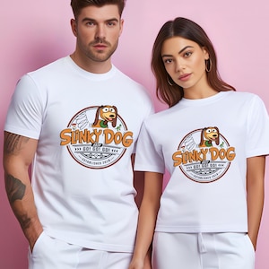 Może przedstawiać: Białe koszulki z okrągłą grafiką Slinky Dog, napisem "Slinky Dog" i "GO! GO! GO!" w kolorze pomarańczowym i brązowym. Projekt zawiera tekst "ESTABLISHED 2018". Koszulki są noszone przez dwie osoby.