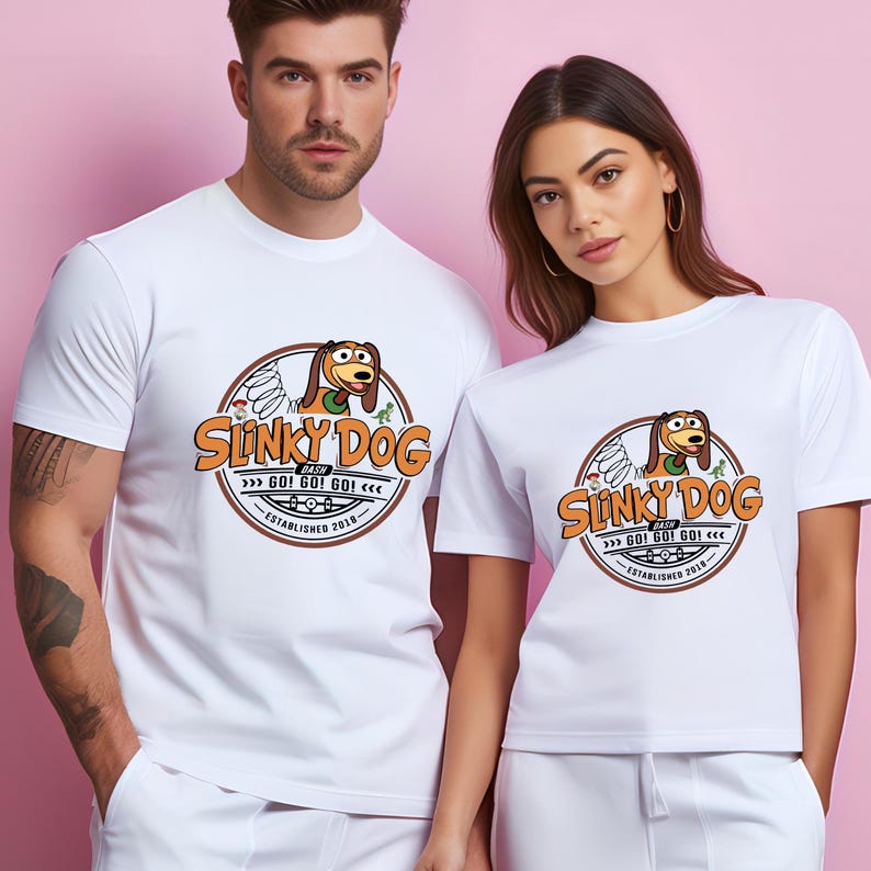 Może przedstawiać: Białe koszulki z okrągłą grafiką Slinky Dog, napisem "Slinky Dog" i "GO! GO! GO!" w kolorze pomarańczowym i brązowym. Projekt zawiera tekst "ESTABLISHED 2018". Koszulki są noszone przez dwie osoby.