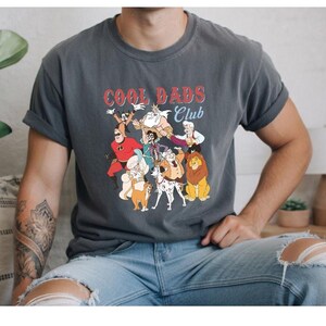 A person wearing a gray t-shirt with a graphic design featuring cartoon characters and the text "Cool Dads Club".