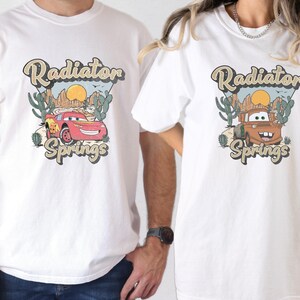 May include: White t-shirts featuring a graphic of a red race car and a brown tow truck with the text "Radiator Springs". The design includes a desert landscape with cacti and a setting sun.
