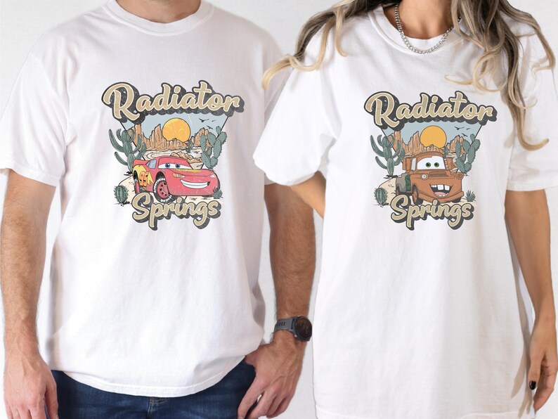 May include: White t-shirts featuring a graphic of a red race car and a brown tow truck with the text "Radiator Springs". The design includes a desert landscape with cacti and a setting sun.