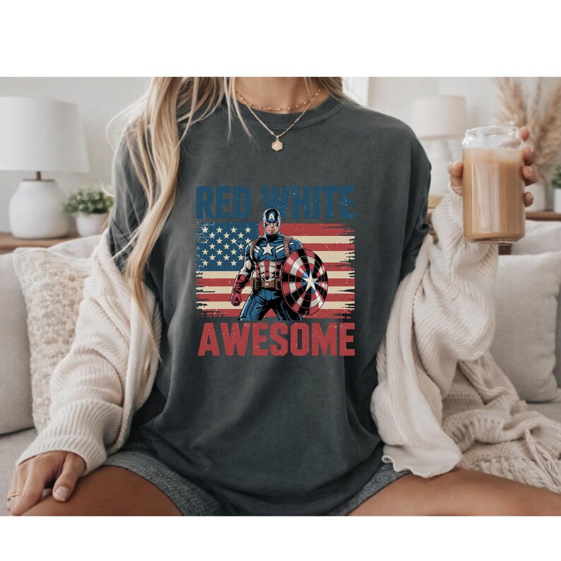 May include: A charcoal gray t-shirt featuring a graphic of Captain America holding a shield in front of a distressed American flag. The text "RED WHITE AND AWESOME" is printed in blue and red block letters above and below the character illustration.