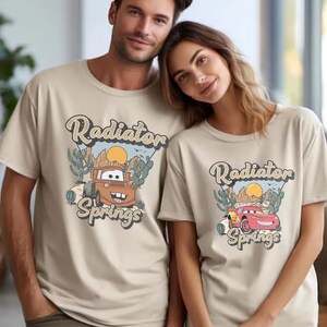 May include: Beige t-shirts featuring a graphic of a cartoon car with the text "Radiator Springs". The design includes a desert landscape with cacti and a sun. The shirts are short-sleeved and worn by two people.