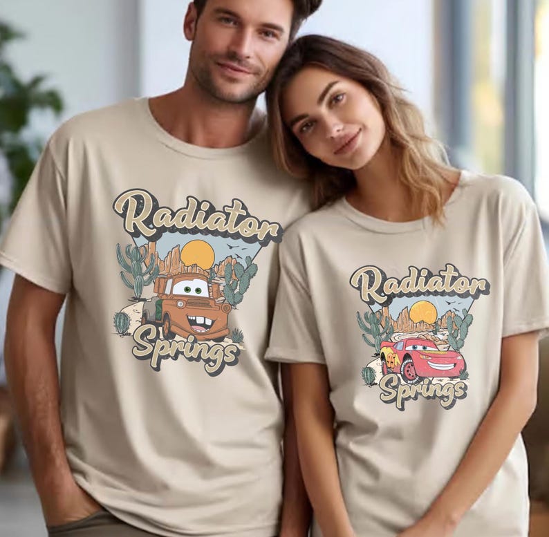 May include: Beige t-shirts featuring a graphic of a cartoon car with the text "Radiator Springs". The design includes a desert landscape with cacti and a sun. The shirts are short-sleeved and worn by two people.