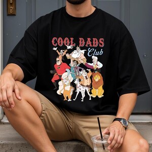 A person wearing a black t-shirt with a graphic of cartoon characters and the text "Cool Dads Club" is sitting on a step.