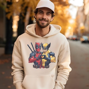 May include: A beige hoodie featuring a graphic of Deadpool and Wolverine forming a heart shape with their hands. The characters are in their signature costumes, with Deadpool in red and Wolverine in yellow and blue.