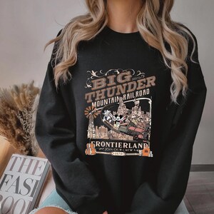 May include: Black sweatshirt featuring a vintage-style graphic for Big Thunder Mountain Railroad. The design includes the text "Big Thunder Mountain Railroad Frontierland Join Our Crew 1979" with cartoon characters and a train.
