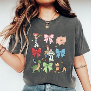 K&ouml;nnte beinhalten: Dunkelgraues T-Shirt mit farbenfrohen Cartoon-Illustrationen von Figuren und Schleifen. Das Design umfasst ein rosa Schwein, einen gr&uuml;nen Dinosaurier und einen Hund mit einem Federk&ouml;rper. Das Shirt ist ein bequemer, l&auml;ssiger Stil.