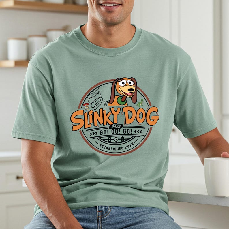 Może przedstawiać: Jasnozielona koszulka z okrągłą grafiką Slinky Dog z Toy Story. Grafika zawiera tekst "Slinky Dog Dash" i "Established 2018". Projekt jest w odcieniach pomarańczu, brązu i zieleni.