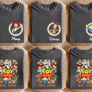 Puede incluir: Camisetas gris oscuro con personajes de Toy Story. Algunas camisetas tienen retratos de personajes con nombres como "Momp", "Daddy" y "Alex". Otras camisetas muestran un grupo de personajes con el texto "TOY STORY Est. 1995".