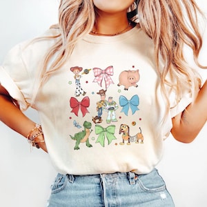 K&ouml;nnte beinhalten: Cremefarbenes T-Shirt mit bunten Cartoon-Figuren und Schleifen. Das Design umfasst Charaktere wie Woody und Buzz Lightyear sowie verschiedene rosa, rote und blaue Schleifen. Das Shirt ist ein Freizeitartikel.