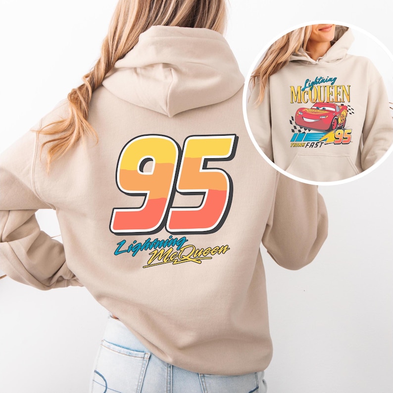 Puede incluir: Sudadera con capucha beige con un gran gr&aacute;fico "95" en amarillo, naranja y rojo, y texto "Lightning McQueen". Una imagen m&aacute;s peque&ntilde;a de Lightning McQueen est&aacute; en la parte delantera. La sudadera tiene una capucha con cord&oacute;n y un bolsillo tipo canguro.