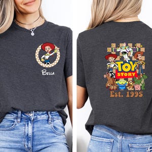 Puede incluir: Camiseta gris oscuro con un gr&aacute;fico del personaje Jessie y el nombre "Bella" en la parte delantera. La parte trasera de la camiseta muestra un gr&aacute;fico de "Toy Story" con personajes y el texto "Est. 1995".